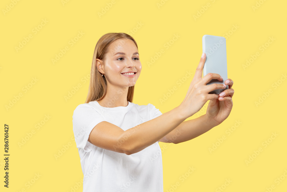 Naklejka premium Caucasian young woman's half-length portrait on yellow studio background. Beautiful female model in white shirt. Concept of human emotions, facial expression. Smiling, making selfie or vlog content.