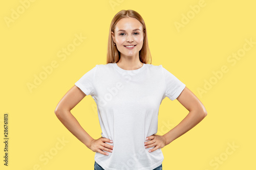 Caucasian young woman's half-length portrait on yellow studio background. Beautiful female model in white shirt. Concept of human emotions, facial expression. Smiling and standing with hands on hips.