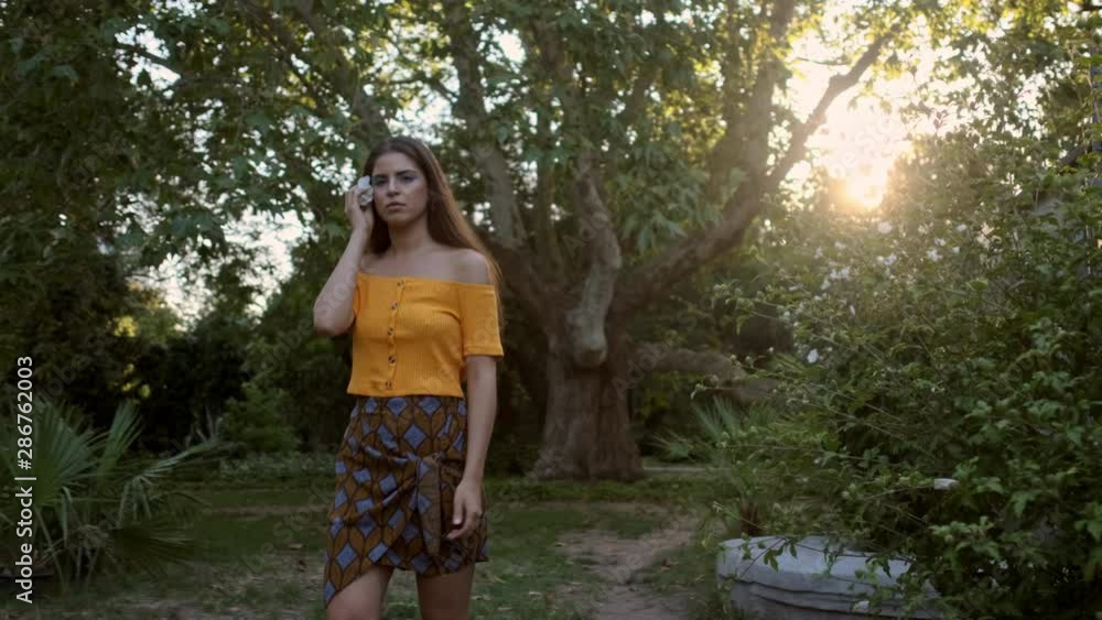 Medium shot of beautiful latin girl passing by putting flower in hair dreamily looking in camera in old garden at sunset Medium shot of beautiful latin girl passing by putting flower in hair dreamily looking in camera in old garden at sunset