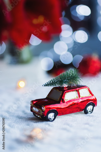 Closeup toy car with Christmas tree on the roof indoor with red home festive decor and blurred bokeh background in daylight.
