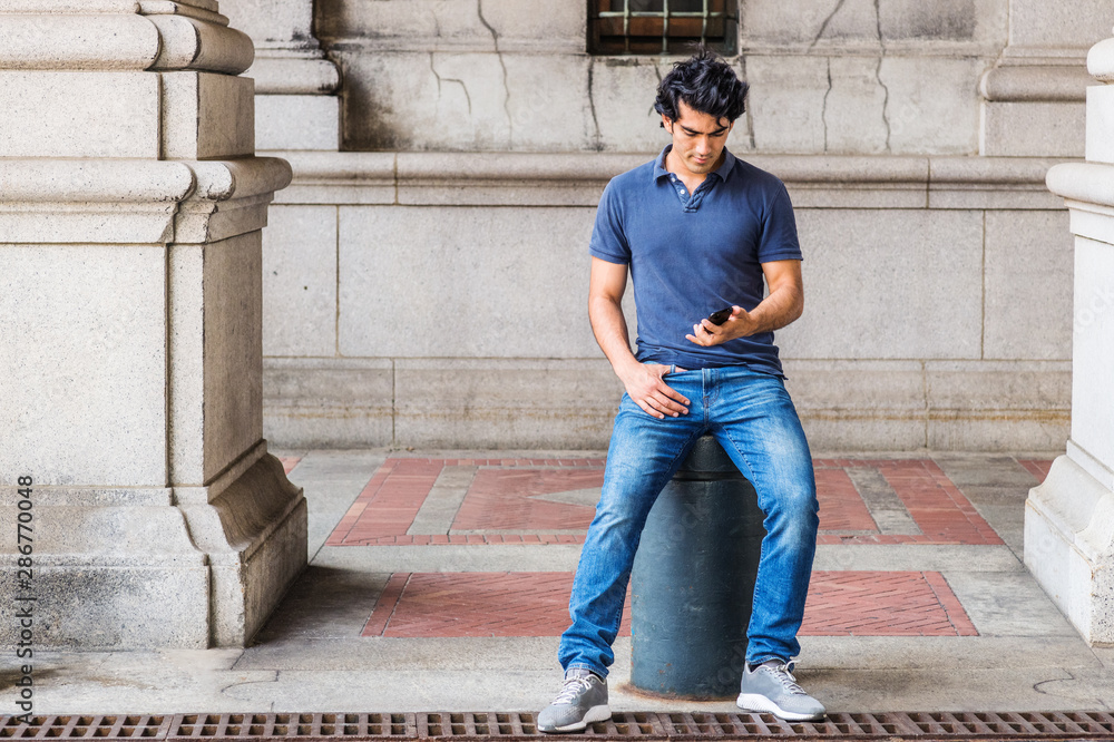 Young Mix Race American college student texting outside, wearing blue short sleeve shirt, jeans, sneakers, sitting on metal pillar on street in school campus in New York City, reading on cell phone.. Young Mix Race American college student texting outside, wearing blue short sleeve shirt, jeans, sneakers, sitting on metal pillar on street in school campus in New York City, reading on cell phone..