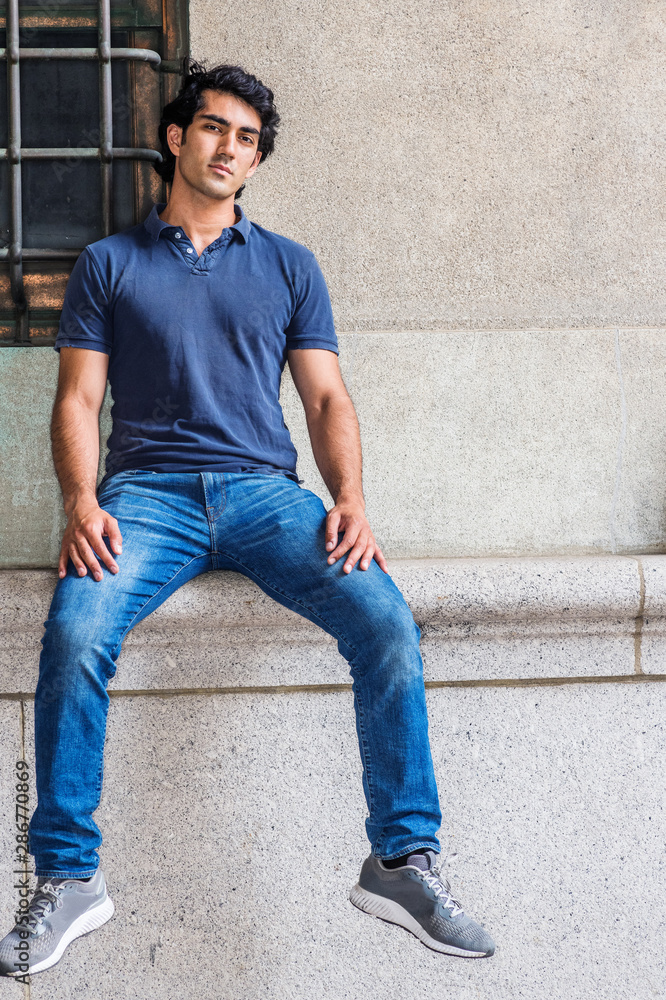 Portrait of Young Mix Race American College Student in New York City, wearing blue short sleeve shirt, blue jeans, sneakers, sitting against old style wall on street in campus, relaxing, thinking.. Portrait of Young Mix Race American College Student in New York City, wearing blue short sleeve shirt, blue jeans, sneakers, sitting against old style wall on street in campus, relaxing, thinking..