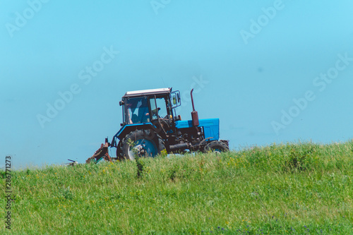 Blue tractor rides on the field against the blue sky and green grass.Beautiful scenery.