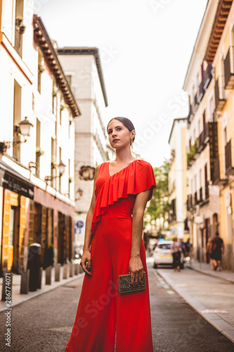 Beautiful brunette young woman with topknot hairstyle wearing red ruffles dress and golden handbag walking on the street. Fashion photo. © EGHStock