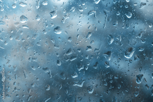 Rain drop on glass pane with blurred autumn dark blue background. Beautiful view from window on rainy day. Water drops on glass with blurred edges.