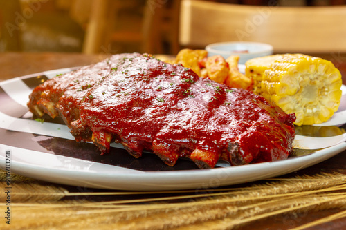 Spicy hot grilled spare ribs from a summer BBQ served with a grilled potato wedges and piece of sweet corn on black and white plate on wooden table.