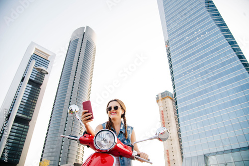 Portrait of a beautiful woman with pigtails smiling on a red motorcycle and towers in the background looking at her phone