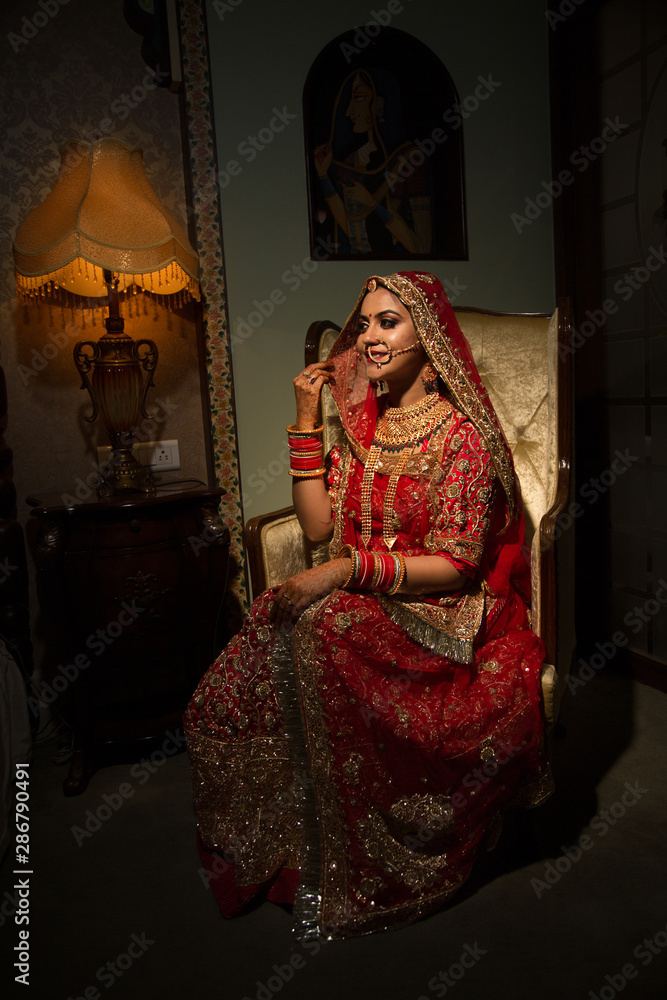 Beautiful traditional Indian girl sitting on sofa like a princess wearing ethnic bridal outfit with heavy makeup and gold jewelery, indian bride portrait