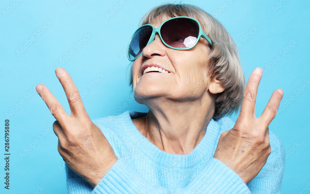 Funny old lady wearing blue sweater, hat and sunglasses showing victory sign Funny old lady wearing blue sweater, hat and sunglasses showing victory sign