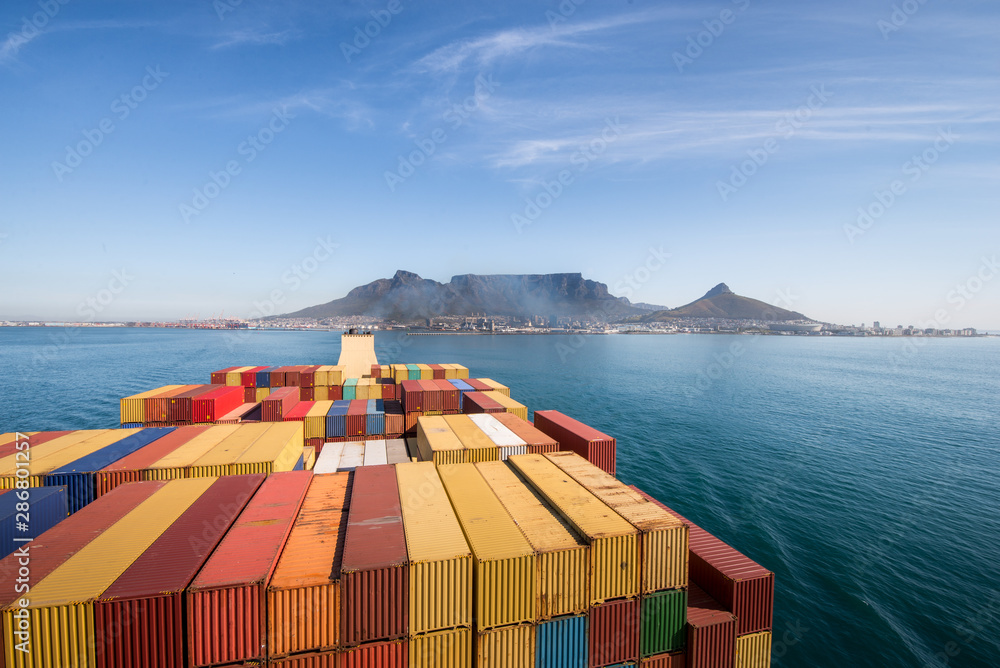 Naklejka premium Duży kontenerowiec opuszczający port w Kapsztadzie z górą stołową i miastem w tle, RPA.
