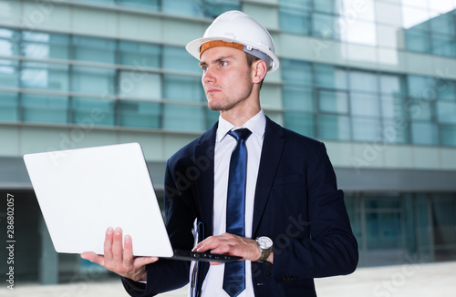 Builder in suit and hat with folder is looking into project in his laptop