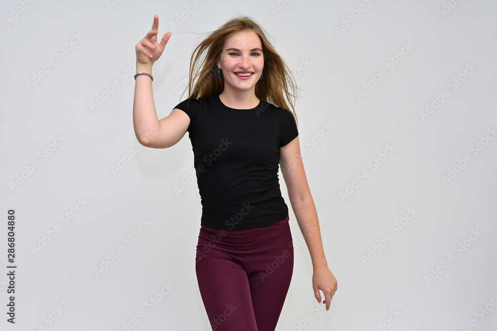A knee-length portrait of a pretty brunette girl with long flowing hair in a black T-shirt and burgundy trousers on a white background. Smiling, showing emotions. A knee-length portrait of a pretty brunette girl with long flowing hair in a black T-shirt and burgundy trousers on a white background. Smiling, showing emotions.