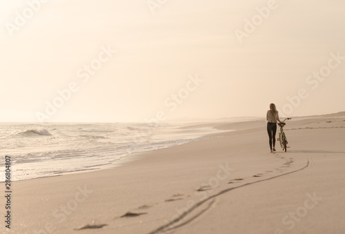 Beautiful blonde woman and bicycle traveling down a quiet beach at sunset.