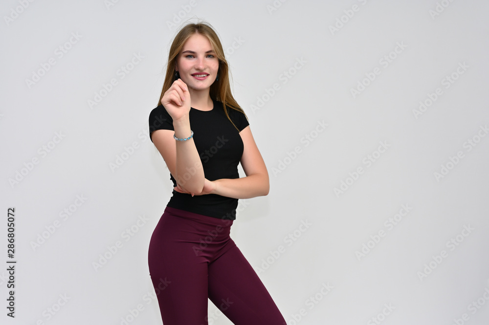 A knee-length portrait of a pretty brunette girl with long flowing hair in a black T-shirt and burgundy trousers on a white background. Smiling, showing emotions. A knee-length portrait of a pretty brunette girl with long flowing hair in a black T-shirt and burgundy trousers on a white background. Smiling, showing emotions.