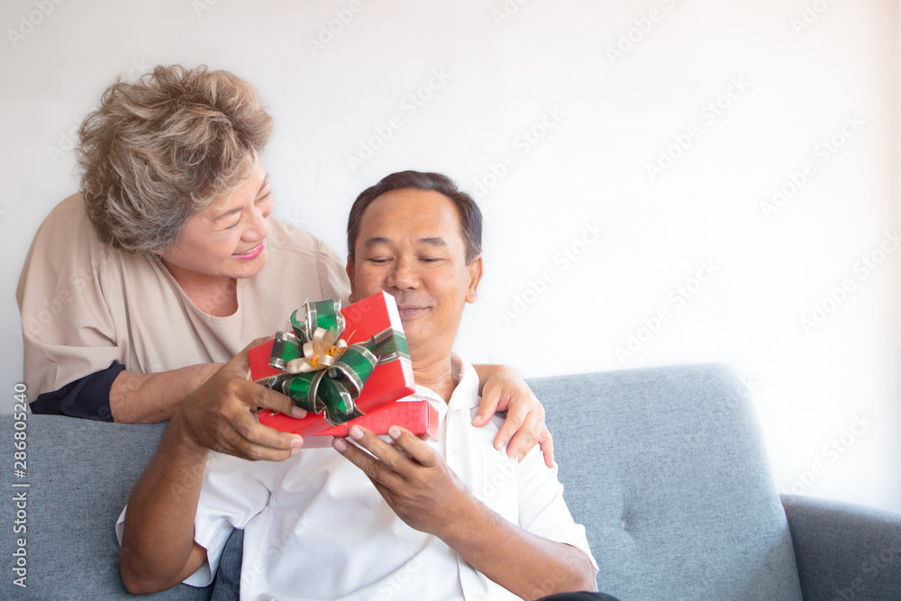 A asian senior couple sitting on couch - wife give a red gift box to husband for present in special day with smile and happy faces. A asian senior couple sitting on couch - wife give a red gift box to husband for present in special day with smile and happy faces.