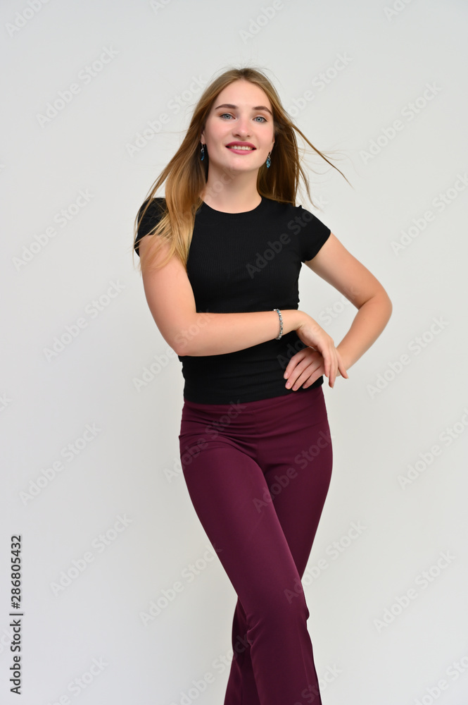 Naklejka premium Full-length vertical portrait of a pretty brunette girl with long flowing hair in a black t-shirt and burgundy trousers on a white background. Smiling, showing emotions.