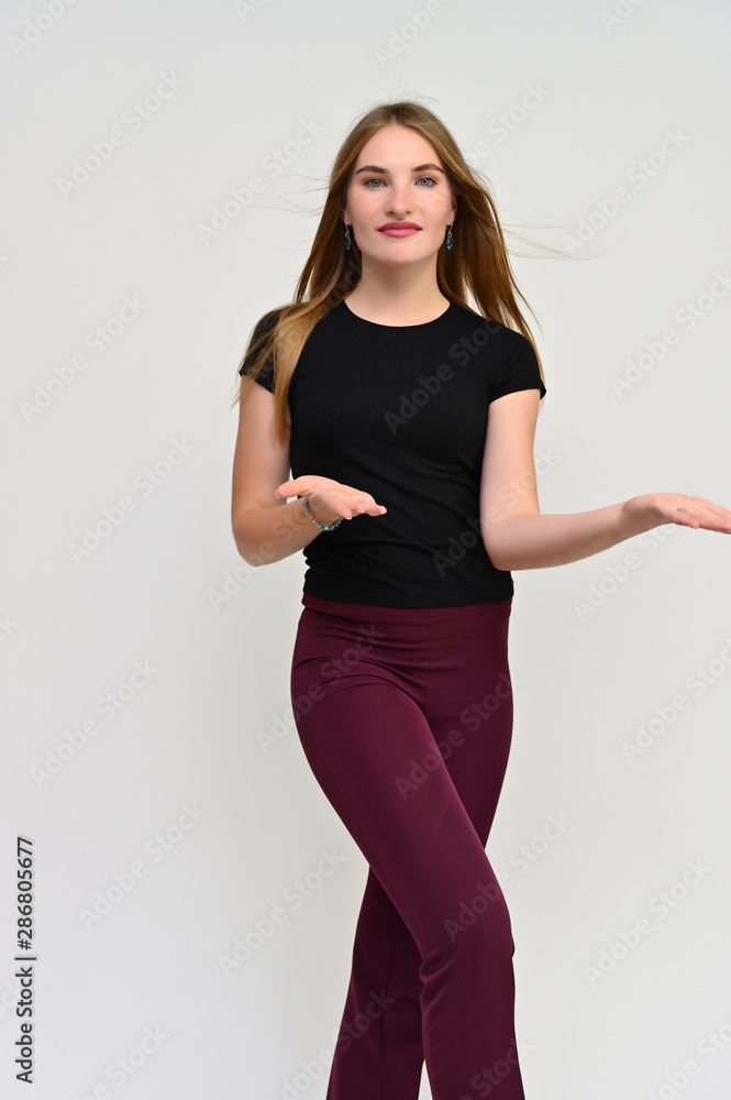 Full-length vertical portrait of a pretty brunette girl with long flowing hair in a black t-shirt and burgundy trousers on a white background. Smiling, showing emotions. Full-length vertical portrait of a pretty brunette girl with long flowing hair in a black t-shirt and burgundy trousers on a white background. Smiling, showing emotions.