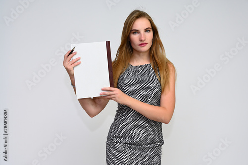 Portrait of a pretty brunette manager girl with long flying hair in a gray dress on a white background with a folder in her hands. Smiling, showing emotions.