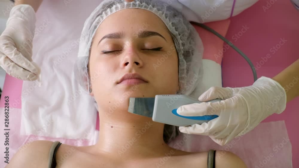 Beauty Specialist Makes Ultrasonic Peeling For Female Client's Face. Cosmetologist doing procedure of cleaning face with ultrasonic scrubber. In beauty salon Beauty Specialist Makes Ultrasonic Peeling For Female Client's Face. Cosmetologist doing procedure of cleaning face with ultrasonic scrubber. In beauty salon