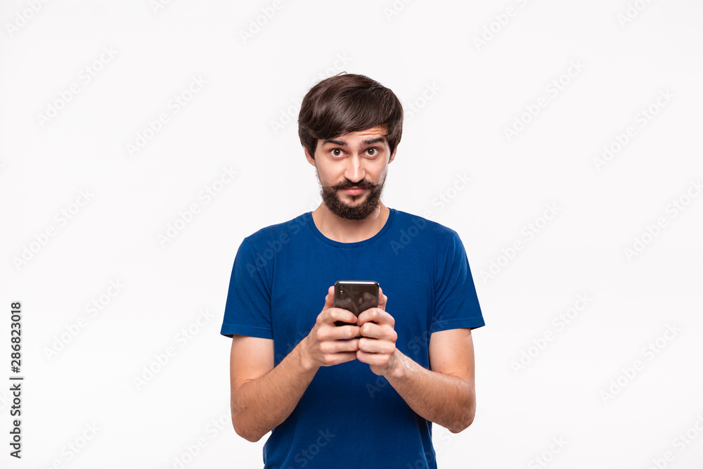 Good looking brunet man in a blue shirt with beard and mostaches shocked holding smartphone looking at the camera standing isolated over white background. Good looking brunet man in a blue shirt with beard and mostaches shocked holding smartphone looking at the camera standing isolated over white background.