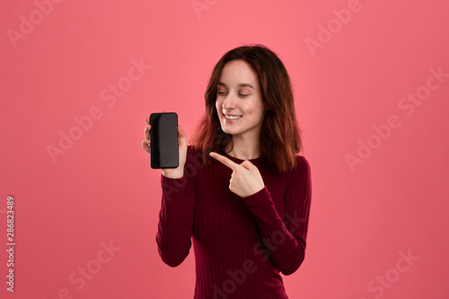 Excited pretty brunette girl pointing at the screen of a mobile phone with a finger while standing on a dark pink background .