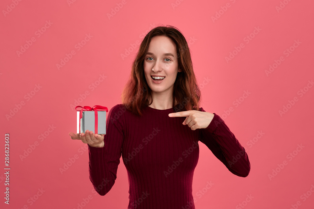 Excited pretty brunette girl holding present box with a ribbon standing isolated on a dark pink background and smiling at the camera. Celebrating special event. Excited pretty brunette girl holding present box with a ribbon standing isolated on a dark pink background and smiling at the camera. Celebrating special event.