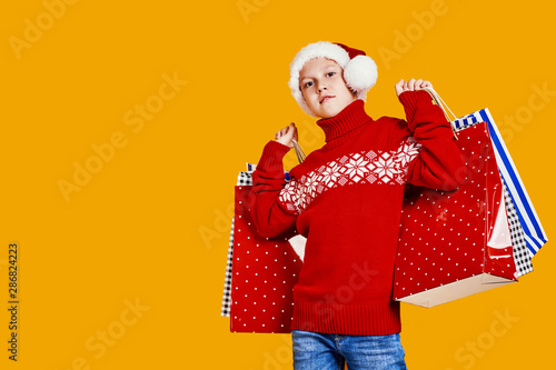 Cute boy in red sweater holding shopping bags with Christmas gifts and looking at camera on yellow background