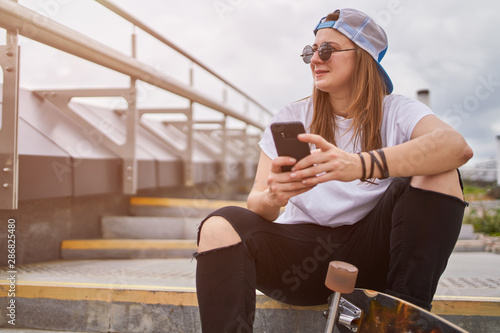Photo of brunette skateboarder in sunglasses with phone in her hands sitting on steps in city