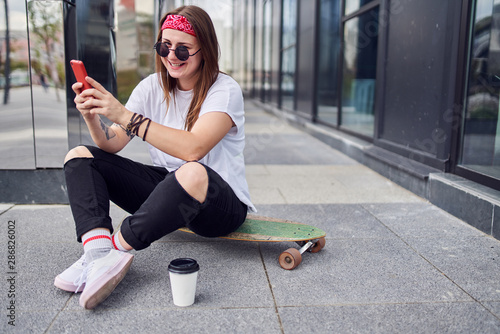 Photo of happy brunette in sunglasses with phone in her hands sitting on skateboard on background of modern buildings in city