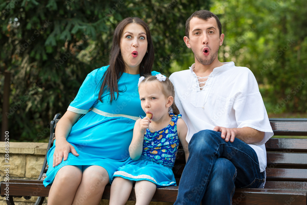 Family sitting on a bench in park. Little girl eating a waffle cup. Woman pregnant. Happy family life concept. Parents grimace. Family sitting on a bench in park. Little girl eating a waffle cup. Woman pregnant. Happy family life concept. Parents grimace.