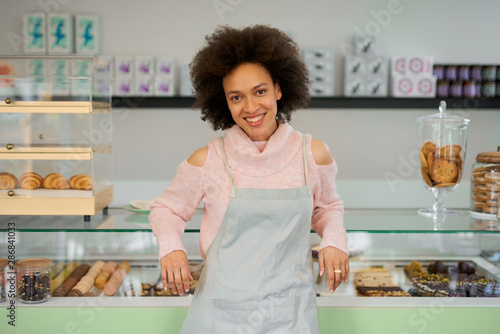 Beautiful smiling mixed race female owner of pastry shop dressed in apron leaning on counter in shop.