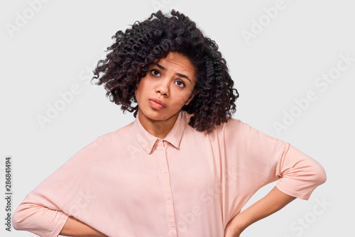 Portrait of questioned Afro young woman looking serious at the camera, during discussing, dressed in pink shirt with curly hair, posing in studio. Student African American girl has confused expression