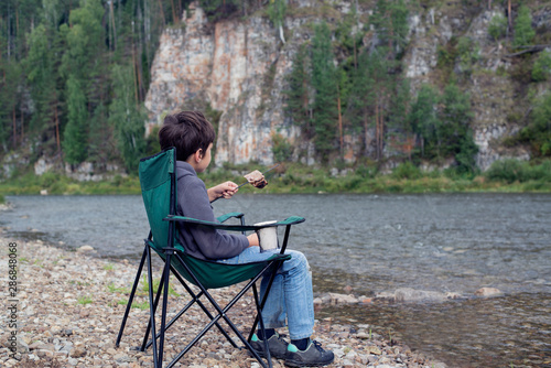 A boy sits on a river bank in a chair with a mug and a piece of fried meat