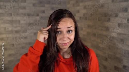A beautiful young woman in a red hoodie twists at her temple on a gray brick background.