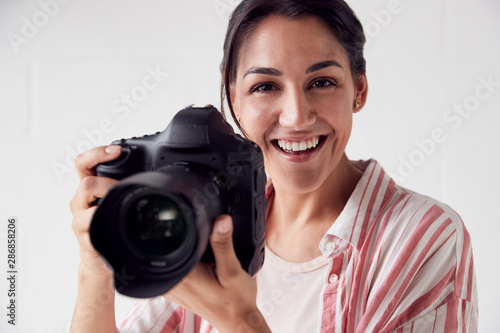 Portrait Of Smiling Female Photographer With Camera Against White Studio Wall