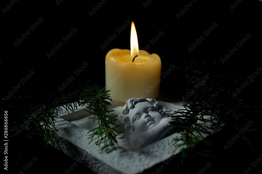 Guardian angel, Fir branches with a burning candle on black background. Christmas tree branches and an Angel lying on the table against darkness. Condolence card concept in christmas time or New year. Guardian angel, Fir branches with a burning candle on black background. Christmas tree branches and an Angel lying on the table against darkness. Condolence card concept in christmas time or New year.