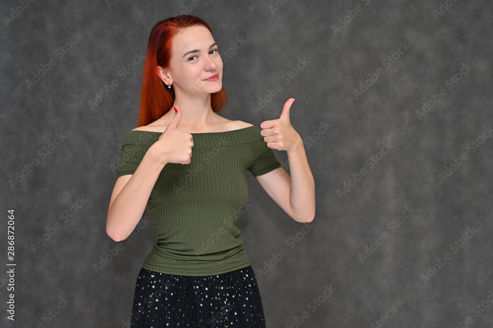 Portrait of a pretty red-haired girl, a happy young woman in a green T-shirt and black skirt on a gray background in the studio. Smiling, showing different emotions.