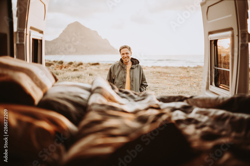 Mann steht vor den offenen Hecktüren eies Camper Vans und blickt in den Van. Im Hintergrund ein Berg und das Meer photo