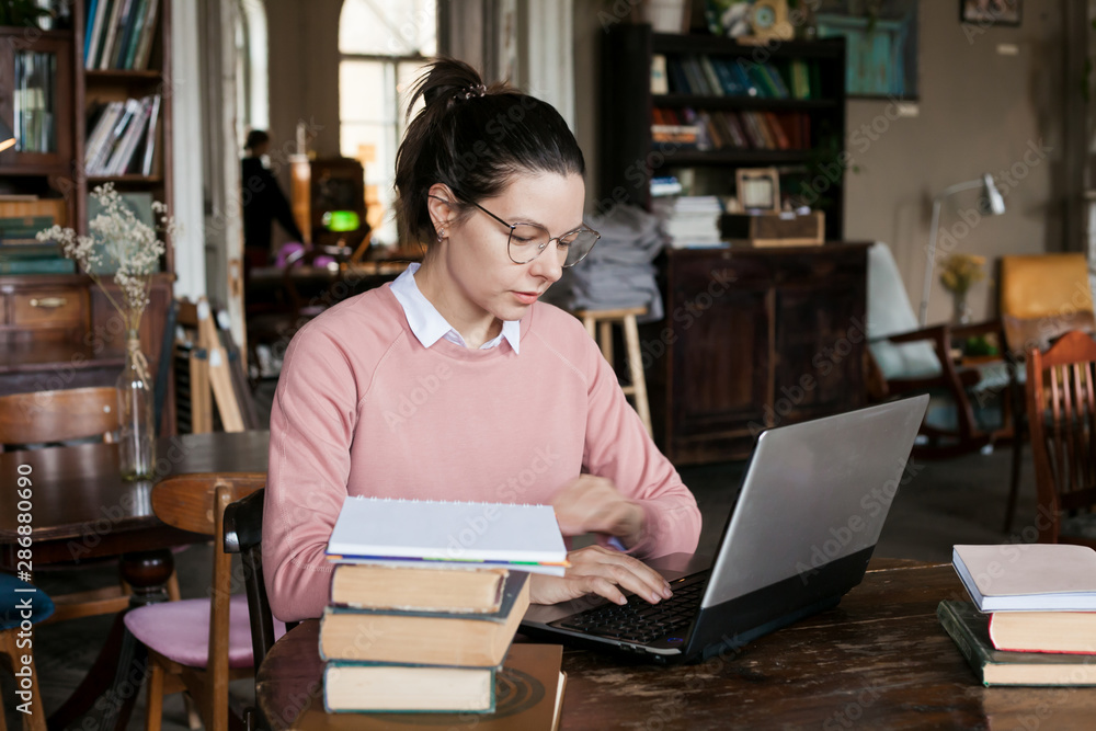 Exam preparation. Attractive brunette student girl in glasses and a pink sweater, with a slight smile sits at a laptop. Exam preparation. Attractive brunette student girl in glasses and a pink sweater, with a slight smile sits at a laptop.