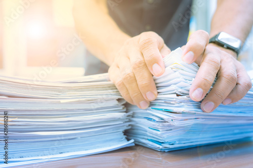 Hands of man searching information from stacks of paper on desk in work office.