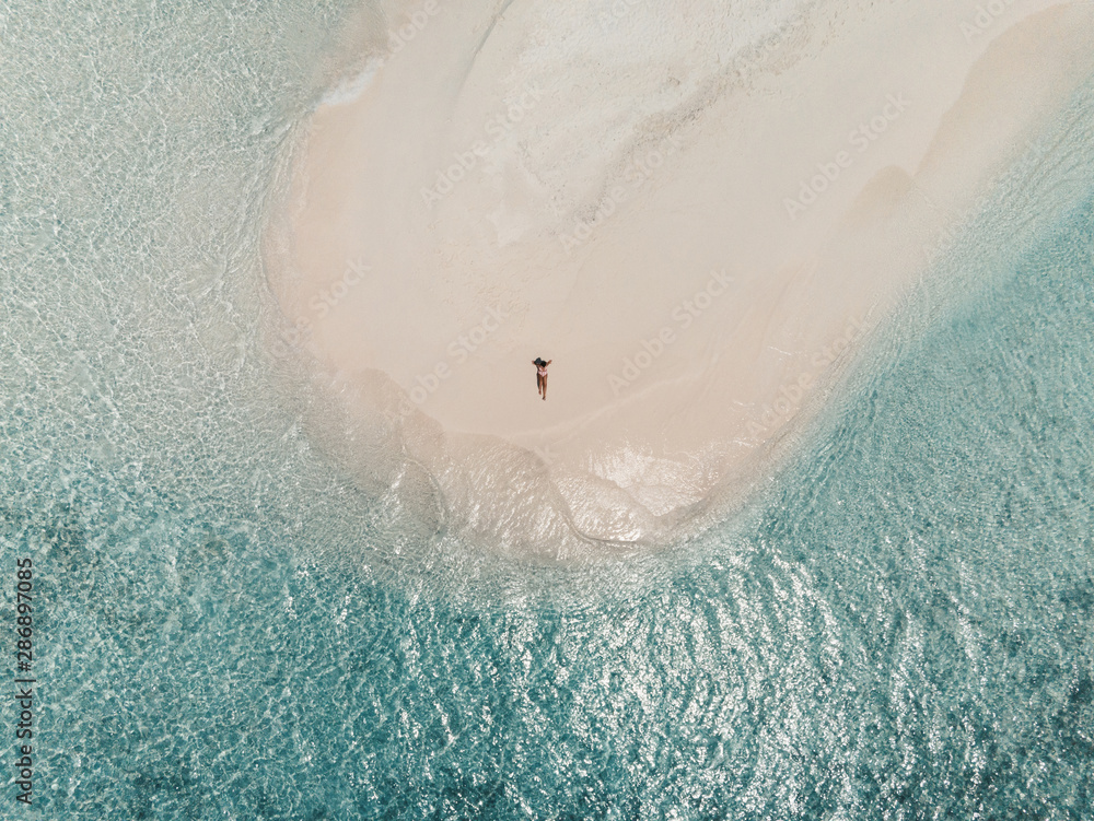 Aerial view of a sandbank in the Maldives sea - A young woman is lying on the white sand while looking at the sea
