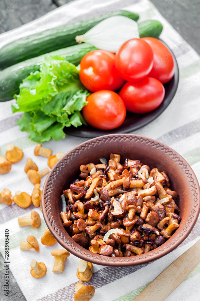 Fried chanterelles with onion in rustic bowl and plate with fresh vegetables for salad on background Fried chanterelles with onion in rustic bowl and plate with fresh vegetables for salad on background