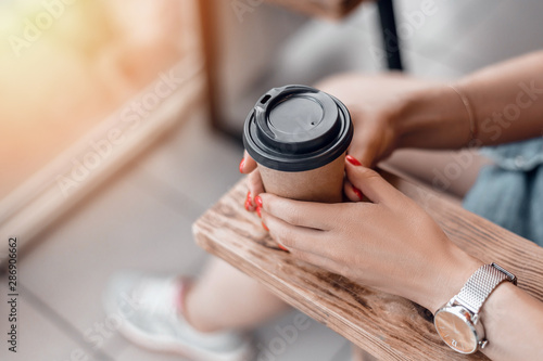 Paper coffee cup to go in woman s hands with red manicure while sitting in cafe.
