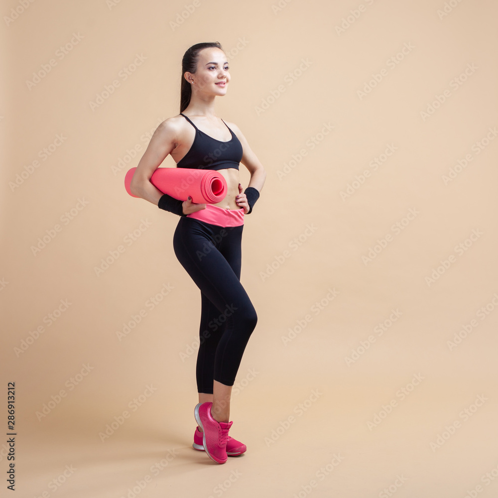 A young girl, a brunette smiling, stands relaxed three-quarters, holding a yoga mat under her arm. A young girl, a brunette smiling, stands relaxed three-quarters, holding a yoga mat under her arm.