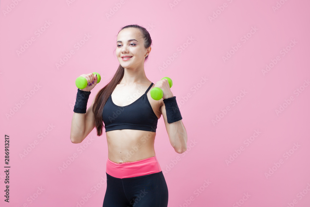 A young athletic woman, with a smile, in black sports top, doing aerobics on pink background with green light dumbbells. A young athletic woman, with a smile, in black sports top, doing aerobics on pink background with green light dumbbells.