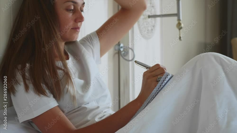 Young woman sitting on bed and writing morning pages to her diary after awake in bedroom. Female writing notes in domestic interior. Young woman sitting on bed and writing morning pages to her diary after awake in bedroom. Female writing notes in domestic interior.