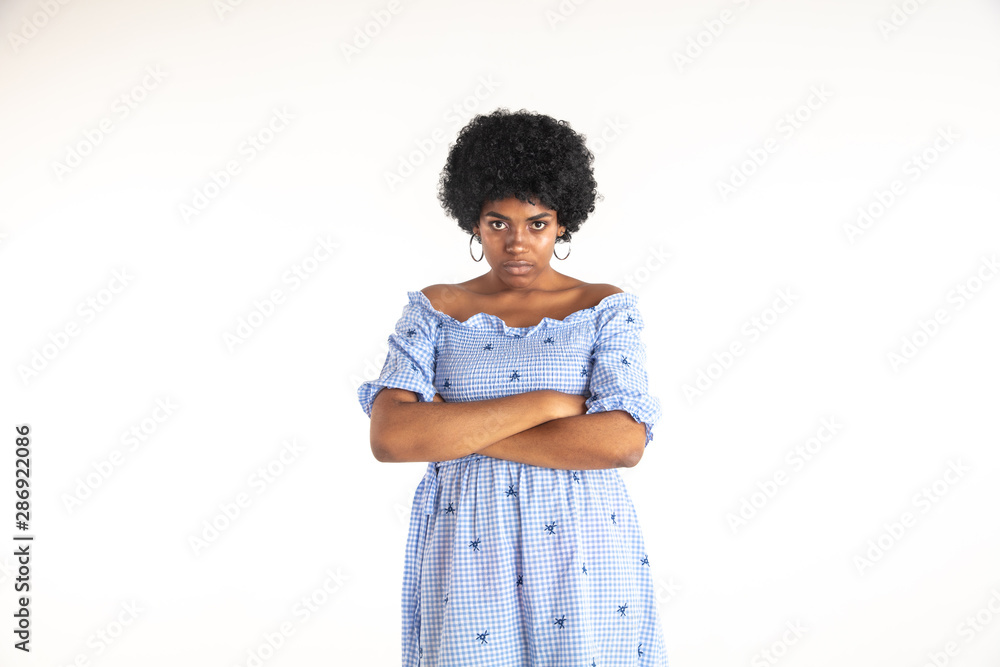 Beautiful female half-length portrait on white studio background. Young emotional african-american woman in blue dress. Facial expression, human emotions concept. Standing with hands crossed