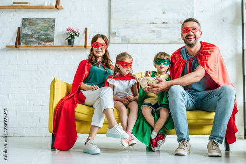 happy family in costumes of superheroes eating popcorn and watching tv at home