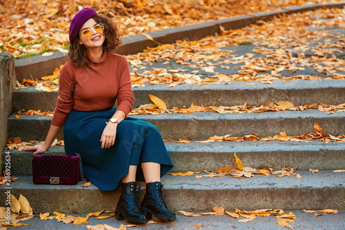 Outdoor autumn portrait of happy smiling lady wearing wrist watch,  beret, orange glasses, turtleneck, blue skirt, black chunky ankle boots, with quilted purple bag, sitting on stairs. Copy space photo