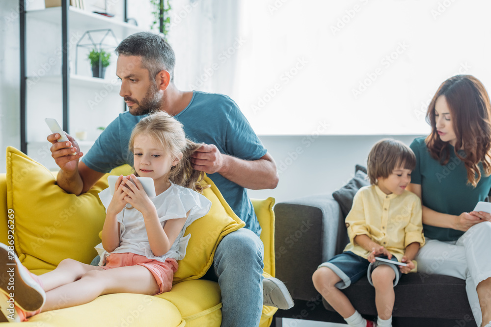 husband and wife with daughter and son sitting on sofa at home and using smartphones husband and wife with daughter and son sitting on sofa at home and using smartphones